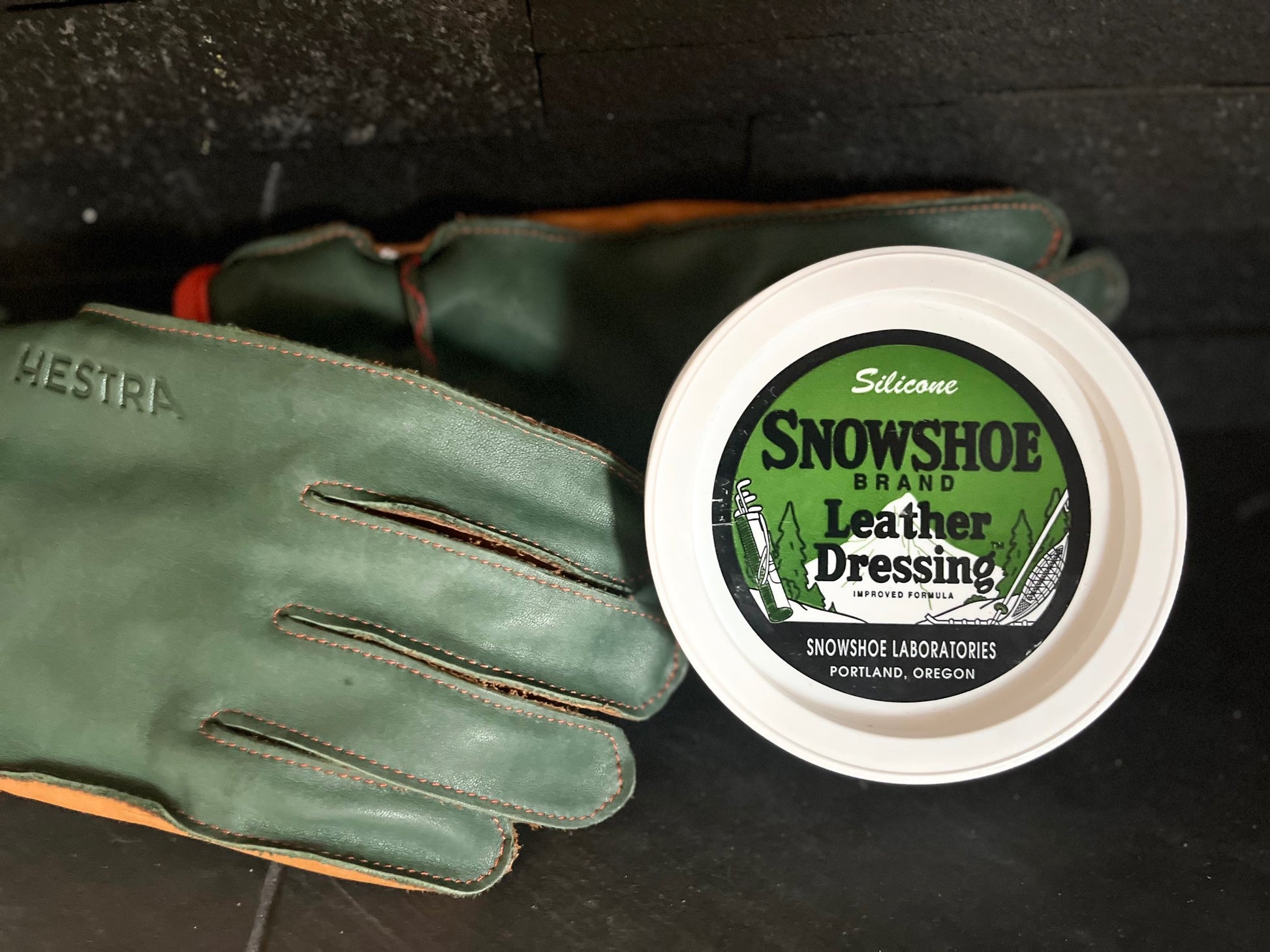 An 8oz tub of Snowshoe Brand Leather Conditioner is sitting next to a green pair of Hestra gloves. 