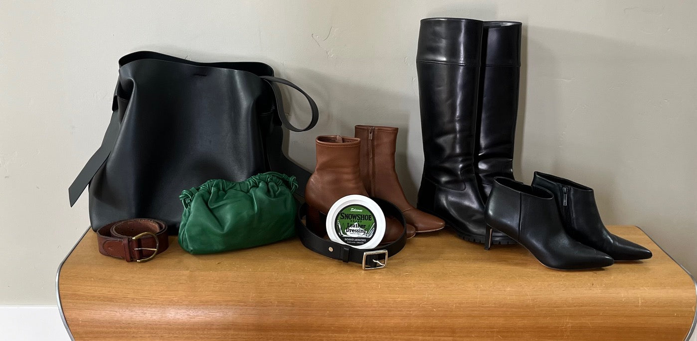 An 8oz tub of Snowshoe Brand Leather Conditioner is displayed on a table with a leather belt, purse, clutch, and three pairs of boots.