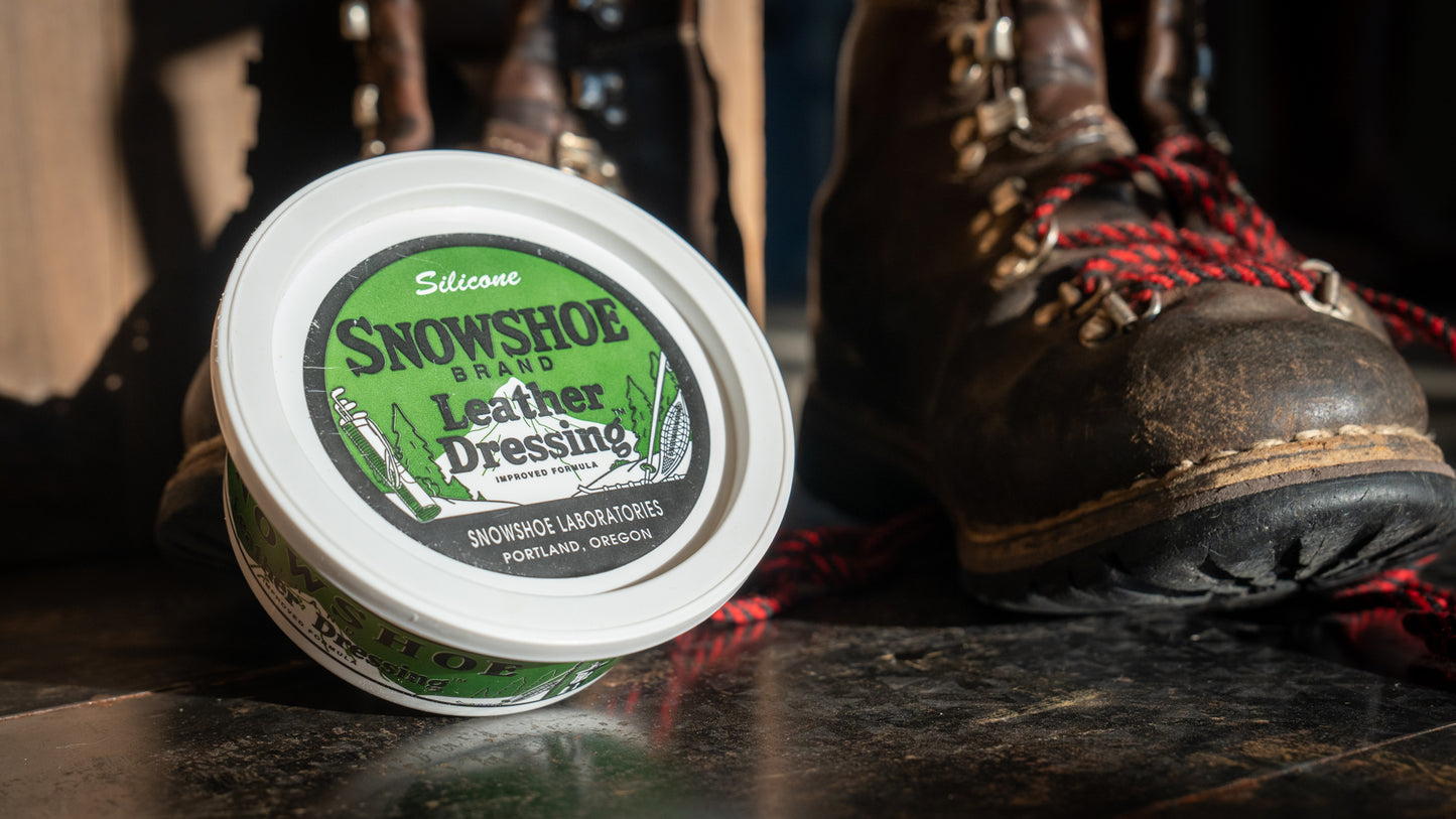 An 8oz tub of Snowshoe Brand Leather Conditioner is propped up against a pair of leather hiking boots. 